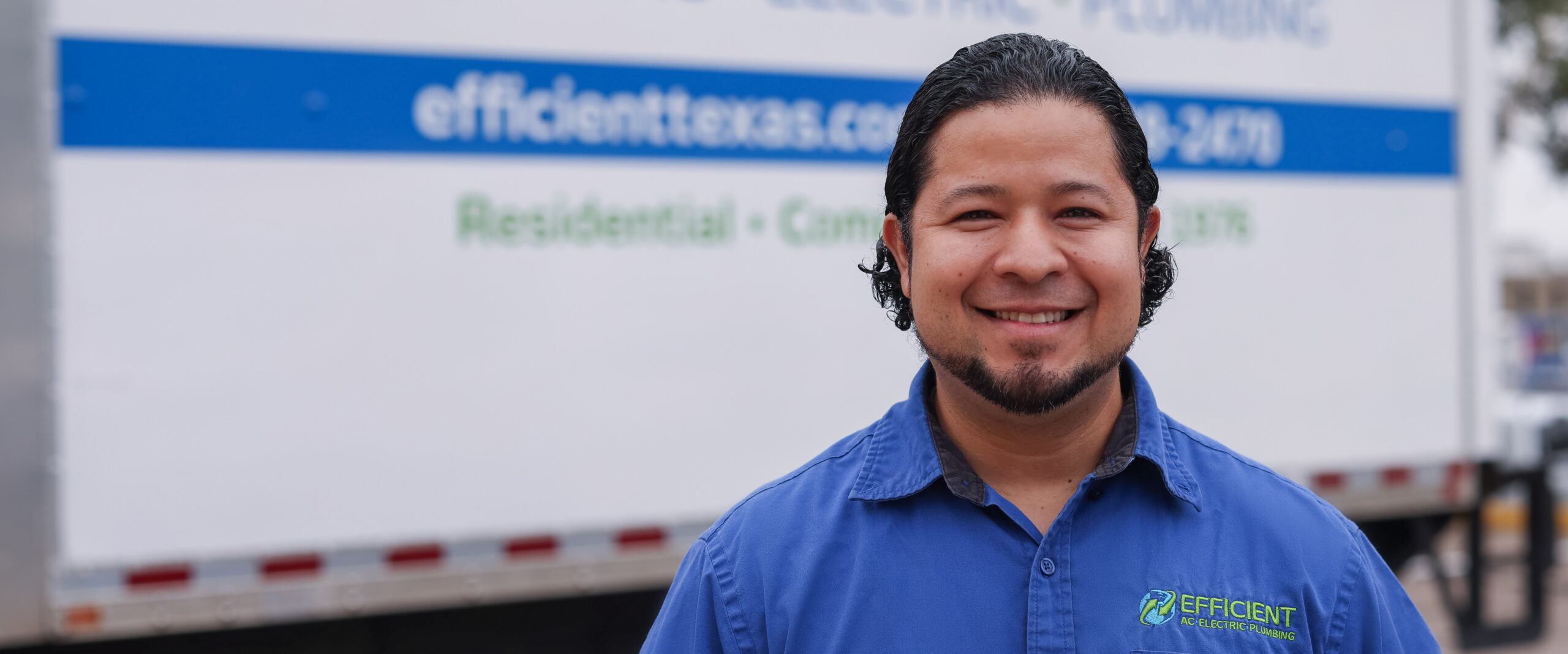 efficient ac electric & plumbing HVAC technician smiling in a blue work shirt standing in front of white work truck in austin tx